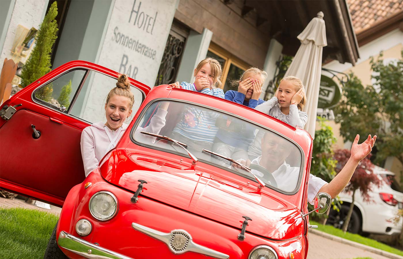 Four children in fun poses on a historic red vehicle in front of the Hotel Alpenhof in Saltusio